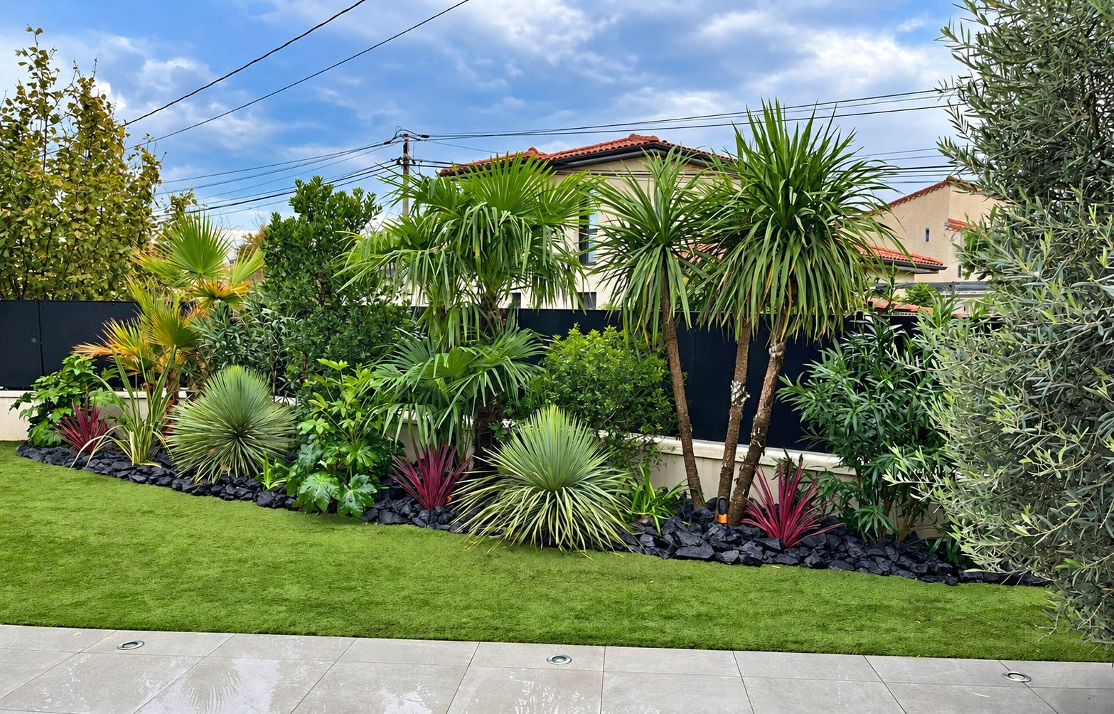 Un jardin aménagé avec un massif de palmiers, de yuccas et de plantes colorées sur un lit de pierres de lave noires, devant une pelouse et une terrasse en carrelage.