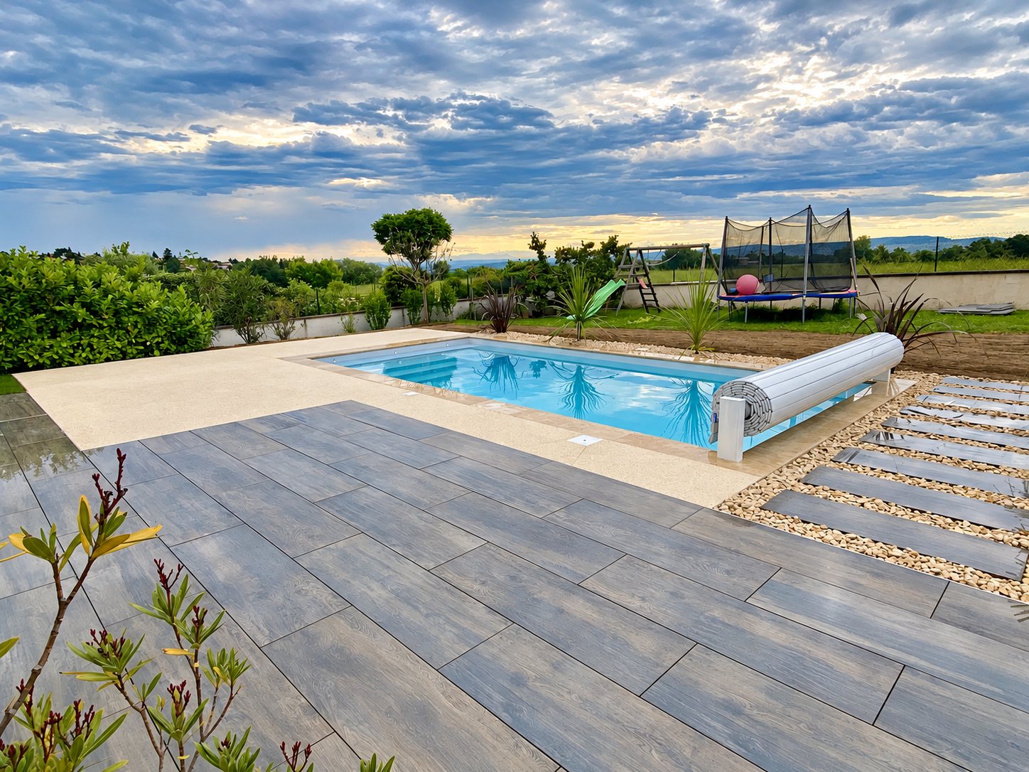 Piscine rectangulaire à l'eau turquoise avec volet roulant enroulé, entourée d'une terrasse en grands carrelages gris et d'un jardin avec trampoline sous un ciel nuageux.