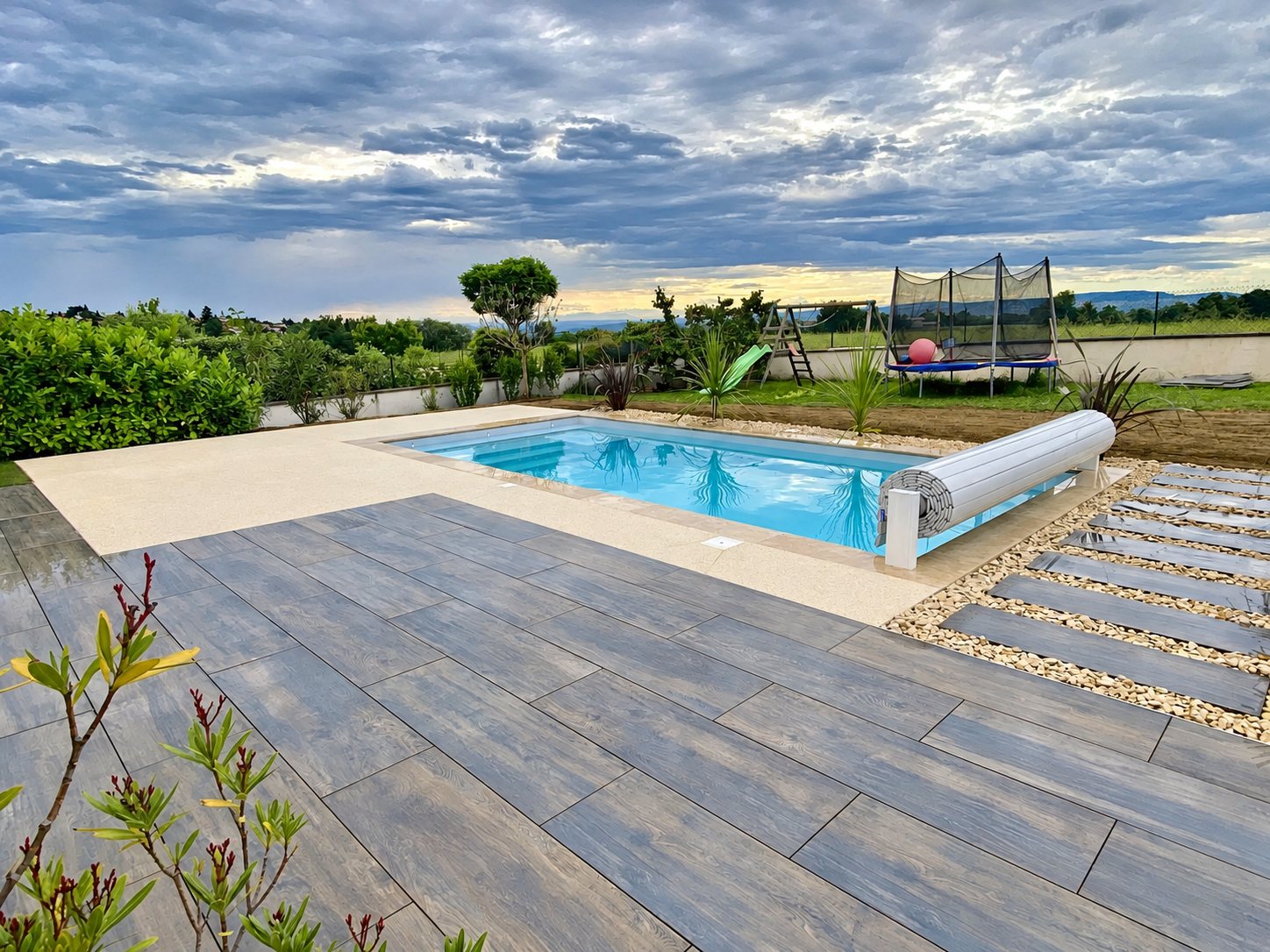 Une piscine rectangulaire à l'eau turquoise, équipée d'un volet roulant, est entourée d'une terrasse en grands carreaux gris effet bois et d'un jardin avec trampoline.