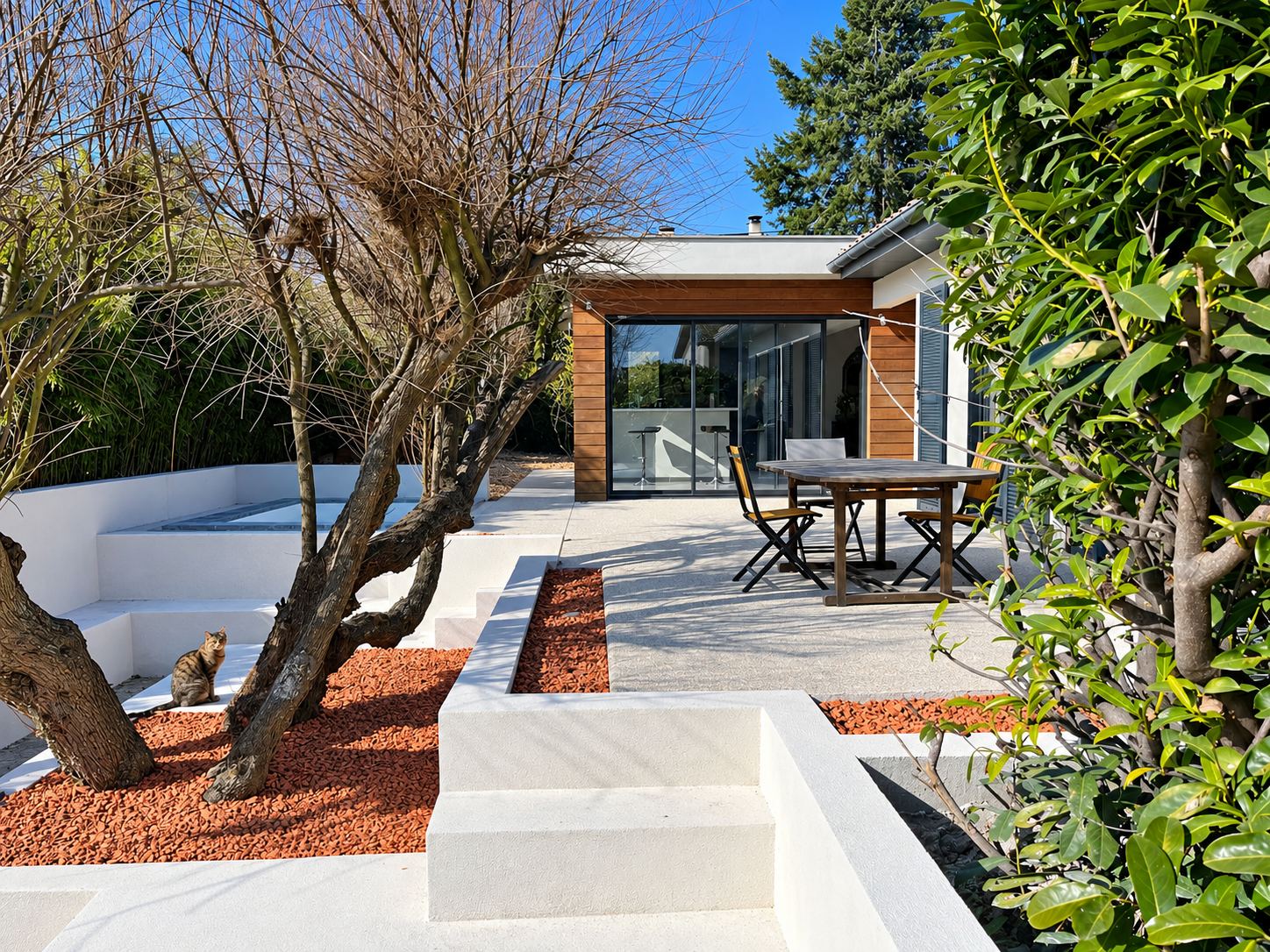 Terrasse en béton clair avec murets blancs, gravier rouge et oliviers taillés devant une maison en bois à grande baie vitrée, où un chat est assis près d'un arbre.