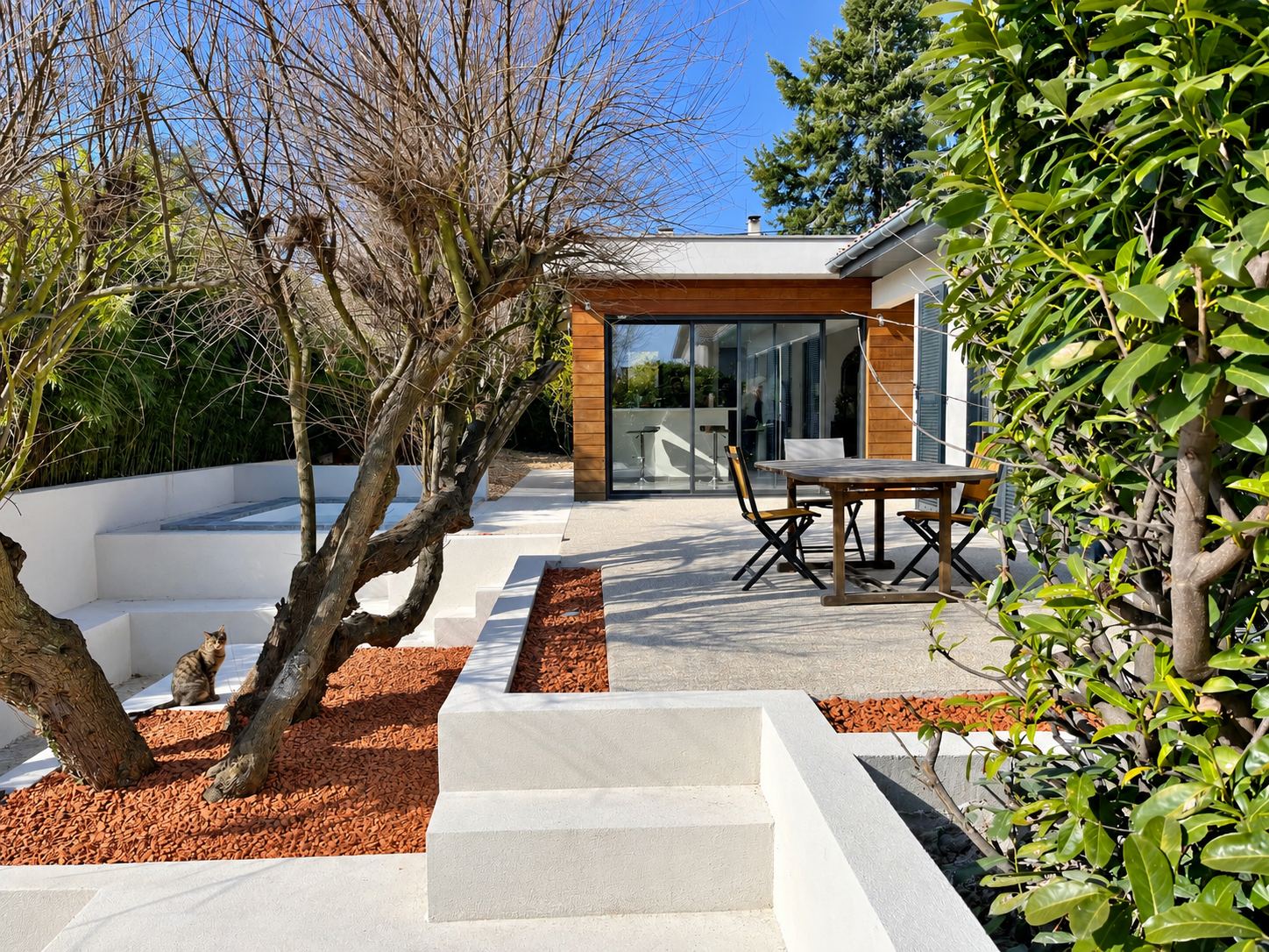 Terrasse en béton clair avec murets blancs, gravier rouge et arbres, devant une maison à bardage bois et grande baie vitrée, où un chat est assis près d'un arbre.