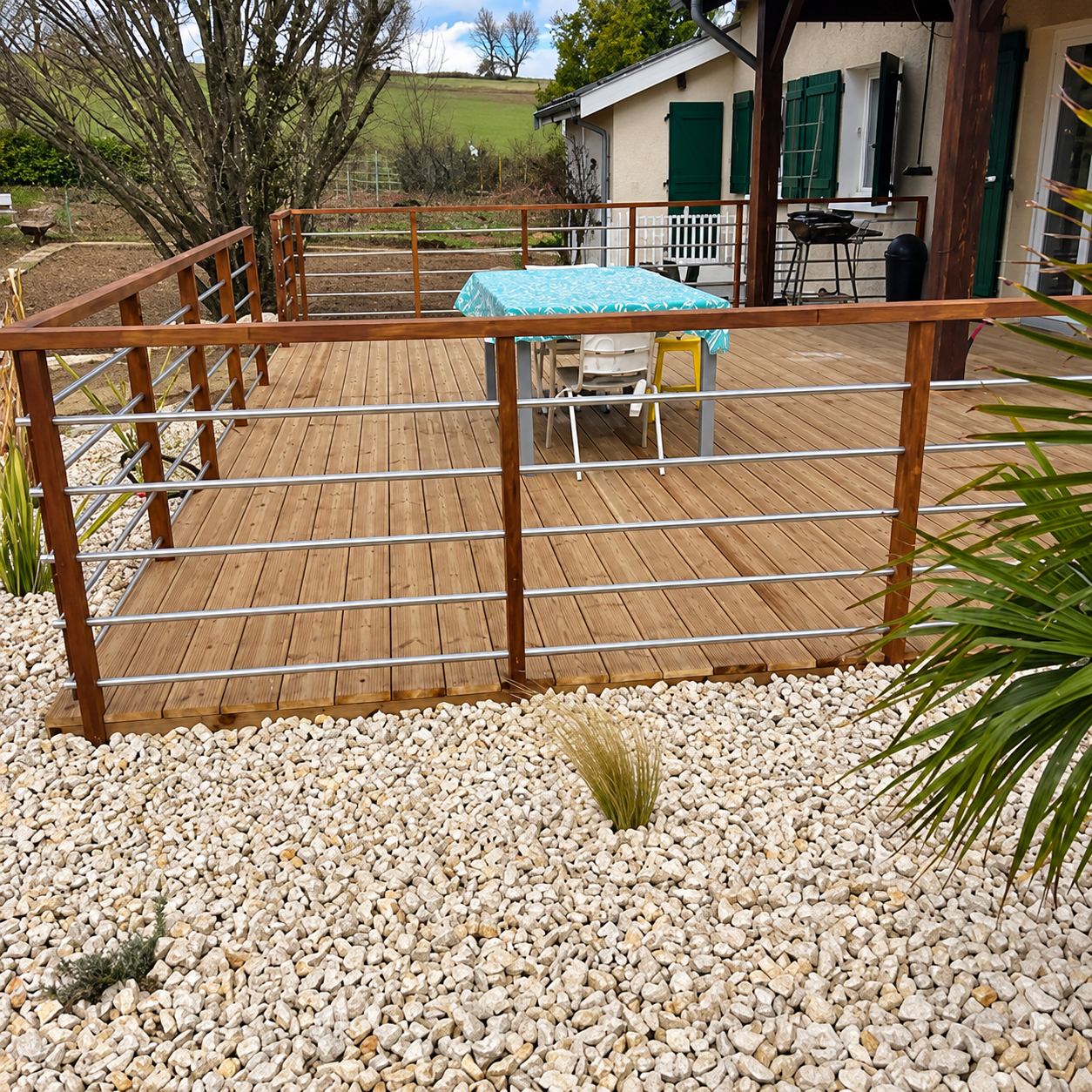 Une terrasse en bois entourée d'un garde-corps à barres horizontales métalliques, bordée de galets blancs, avec une table bleue et une maison aux volets verts en arrière-plan.
