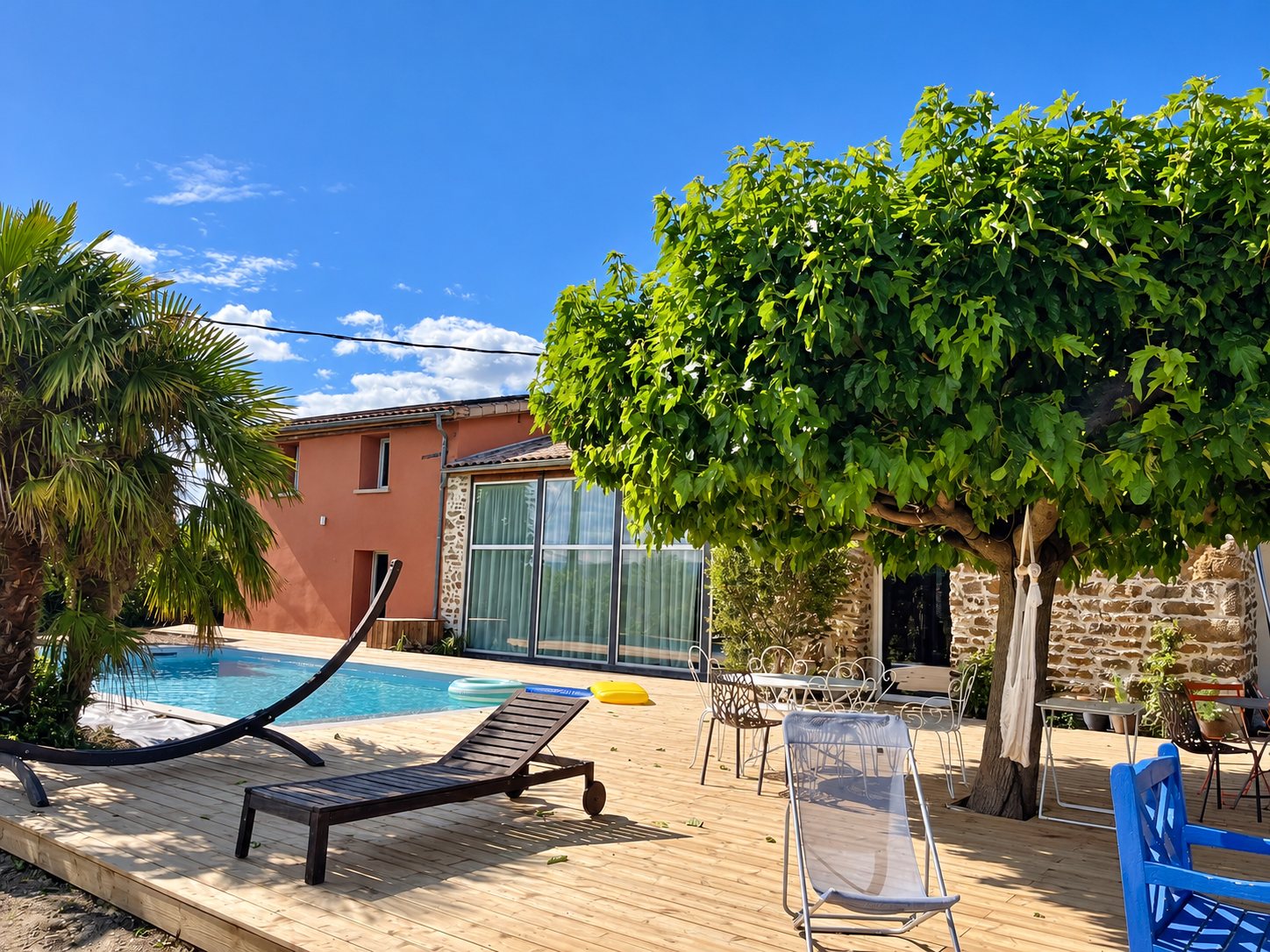 Une terrasse en bois ensoleillée entourée d'une piscine, d'un platane, de chaises et d'une maison rose avec grande baie vitrée.