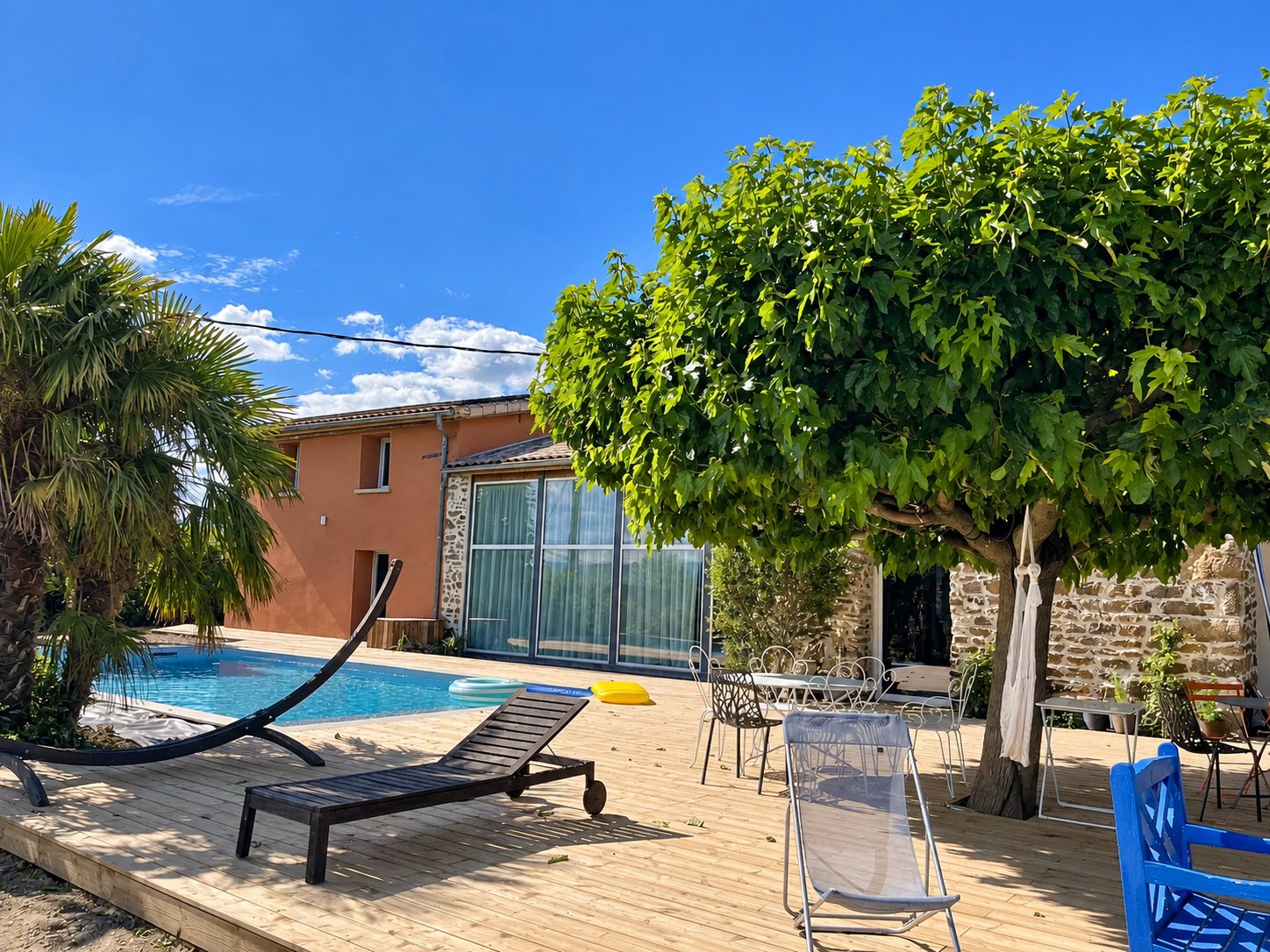 Une terrasse en bois ensoleillée entourée d'une piscine, d'un platane, de chaises et d'une maison en pierre et crépi rose.