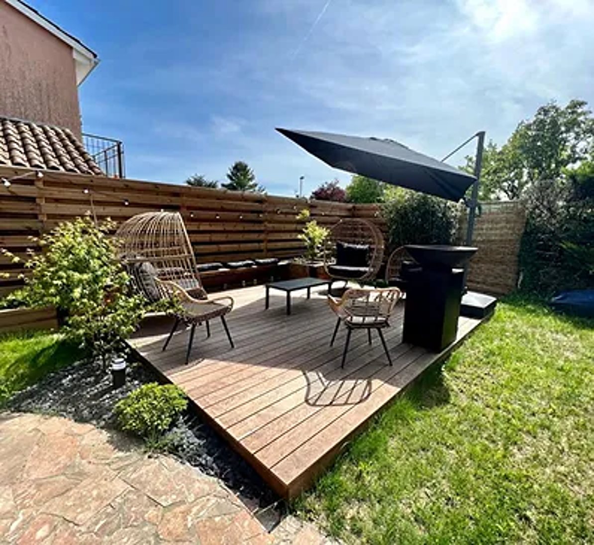 Une terrasse en bois surélevée avec fauteuils en rotin, table basse et grand parasol noir, entourée d'une clôture en bois et de pelouse.